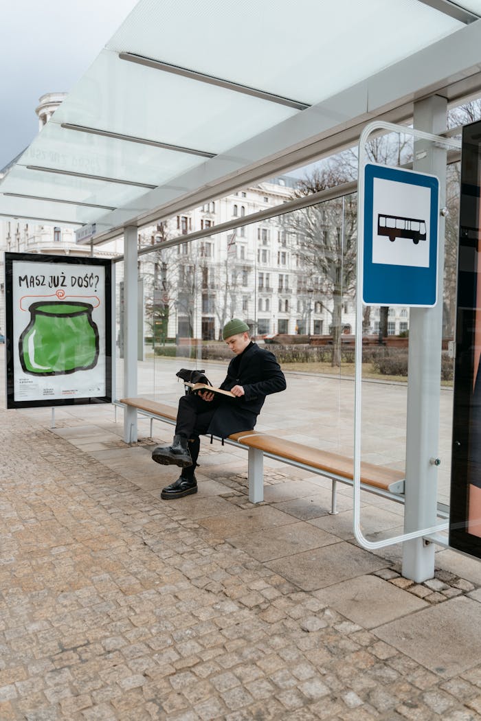 contact-img Man in coat reading a book while sitting at an outdoor bus stop on a cobbled street.